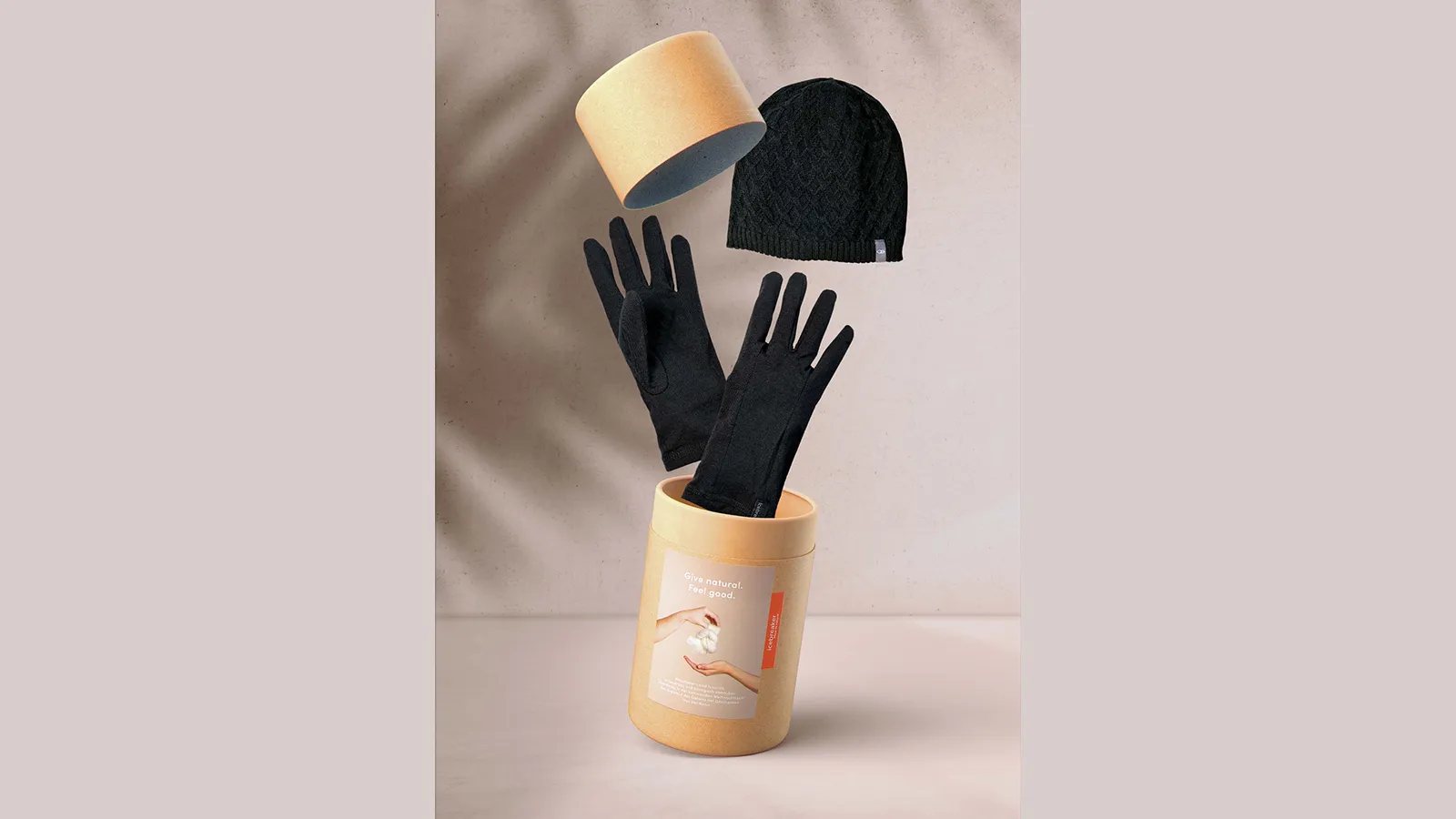 Runde Icebreaker-Verpackung aus Kraftpapier mit Aufschrift Give natural, Feel good: Deckel schwebt ab, darüber eine schwarze Strickmütze, schwarze Handschuhe ragen aus der offenen Dose