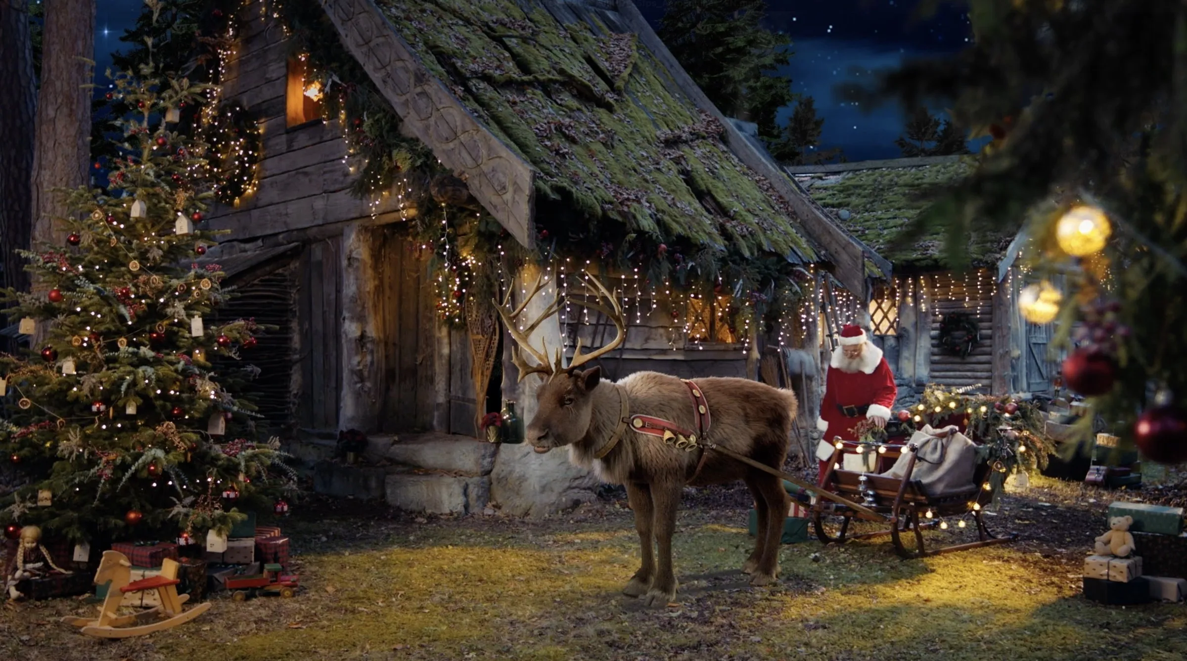 Rentier mit Geweih zieht einen Schlitten vor einer beleuchteten Holzhütte, ein Weihnachtsmann steht daneben, Geschenke und ein geschmückter Christbaum im Vordergrund