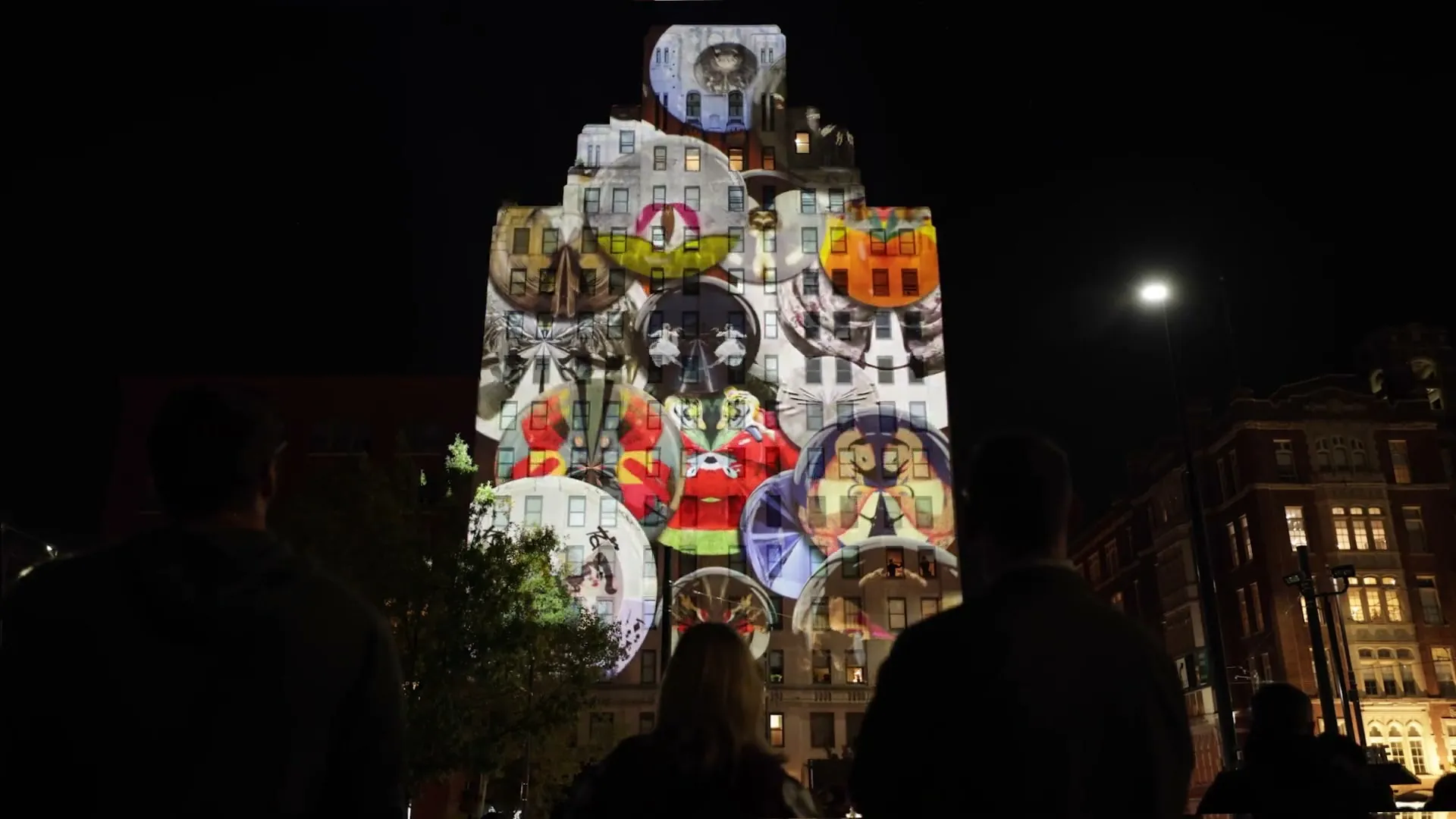 Projektion auf dem Hochhaus bei Nacht, im Vordergrund die Silhouetten von Zuschauern