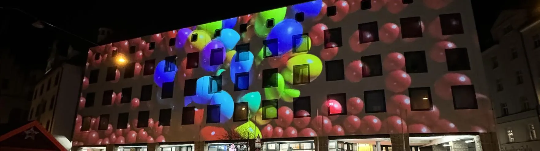 Fassadenprojektion mit rosa Kugelmuster und bunten Ballonformen in Blau und Grün auf einer Häuserfront, eine Straßenlaterne davor