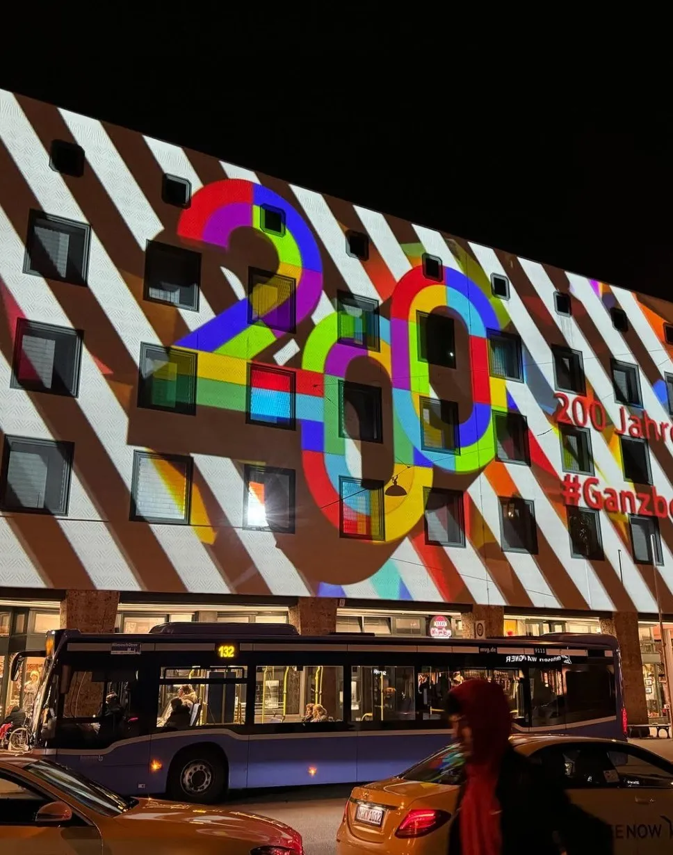 Fassadenprojektion mit der Zahl 200 in Regenbogenfarben auf gestreifter Fassade, davor ein Linienbus und parkende Autos an einer Straße