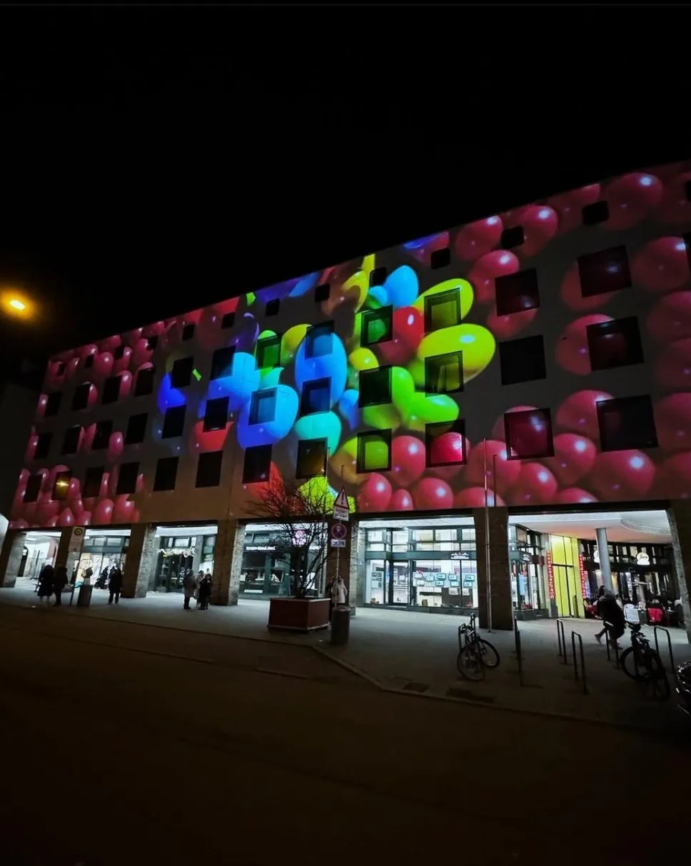 Fassadenprojektion mit bunten ballonartigen Kugeln in Rosa, Blau und Grün auf einer Häuserfront, Fußgänger und Fahrräder davor