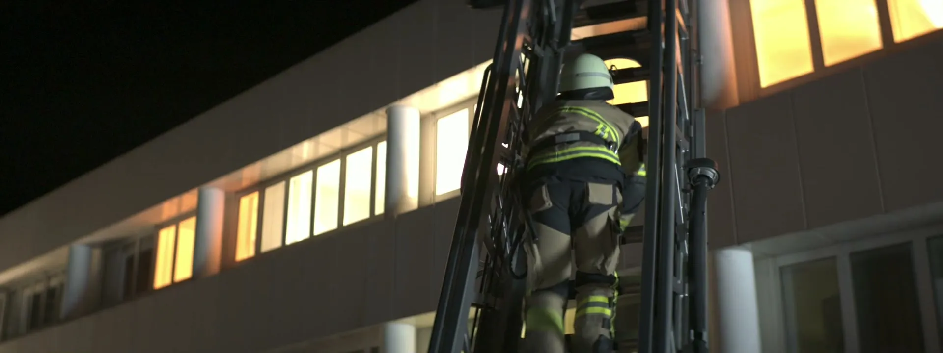 Feuerwehrmann in Einsatzkleidung steigt eine Leiter hinauf, daneben ein nachts beleuchtetes Bürogebäude mit hell erleuchteten Fenstern: die Originalaufnahme